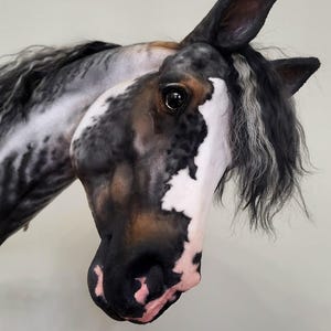 Peut inclure: Une sculpture détaillée d'une tête de cheval, avec un mélange saisissant de couleurs noir, blanc et marron. Le cheval a une longue crinière fluide avec des mèches grises et noires. L'œil de la sculpture est détaillé et le nez est un mélange de rose et de noir.