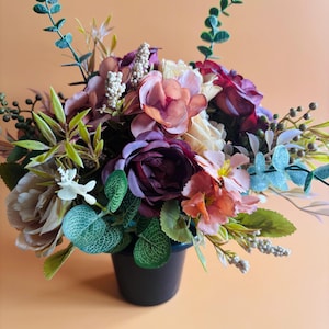 May include: An artificial floral arrangement in a black pot featuring a mix of deep purple, dusty pink, and cream-coloured flowers. The bouquet includes various green eucalyptus-style leaves, small white berry sprigs, and slender stems.