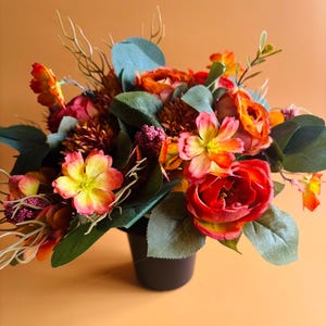 May include: A floral arrangement in a dark container featuring a mix of artificial flowers in shades of orange, red, and yellow, accented with green eucalyptus leaves, small purple berry clusters, and thin, light-coloured wispy stems.