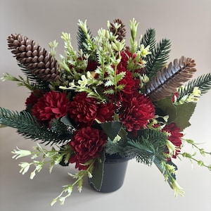 May include: A floral arrangement in a black pot featuring deep red chrysanthemum-like flowers, brown pinecones, green pine needles, and small pale yellow sprigs. The composition is dense and rounded, combining dark foliage with bright accents.