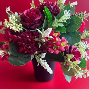 May include: A floral arrangement in a dark container featuring deep red roses, dark red carnations, and smaller bright red blossoms. The bouquet includes green leaves and clusters of small white flowers, creating a dense, multi-textured display.