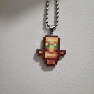 Op de afbeelding: Een pixelachtige hangerketting met een bruin en beige totemontwerp met groene ogen. De hanger is bevestigd aan een zilveren kogelketting. De ketting wordt gepresenteerd tegen een witte achtergrond.