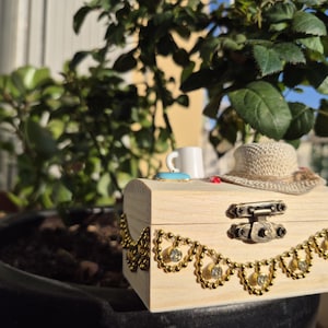 May include: A small, light-colored wooden box with a decorative gold chain and clasp. A miniature white mug, a blue coaster, and a woven hat rest on top. The box is set in front of a lush green plant.
