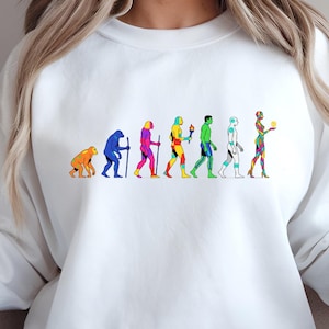 May include: White sweatshirt featuring a colorful graphic of human evolution. The design shows a progression of figures, each in a different color, from a primate to a modern human. The sweatshirt is a casual, comfortable style.