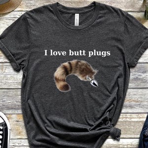 May include: Dark gray t-shirt with the text "I love butt plugs" above an illustration of a furry tail with a metal plug. The shirt is displayed with black sneakers, glasses, a watch, and jeans.