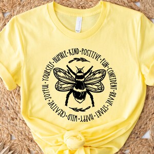 May include: A yellow t-shirt featuring a black and white bee design. The bee is surrounded by a circular text that includes words like "joyful", "kind", and "brave". The shirt is made of a soft material.