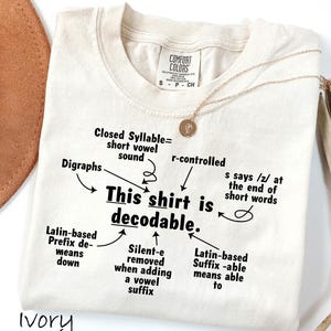 May include: An ivory t-shirt featuring black text that reads, This shirt is decodable, surrounded by arrows pointing to linguistic terms including Closed Syllable, Digraphs, r-controlled, Latin-based Prefix, Silent-e, and Latin-based Suffix.