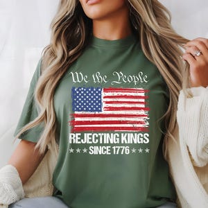 May include: Olive green t-shirt featuring a distressed American flag graphic with the text "We the People REJECTING KINGS SINCE 1776". The design includes red and white stripes, blue stars, and white text.