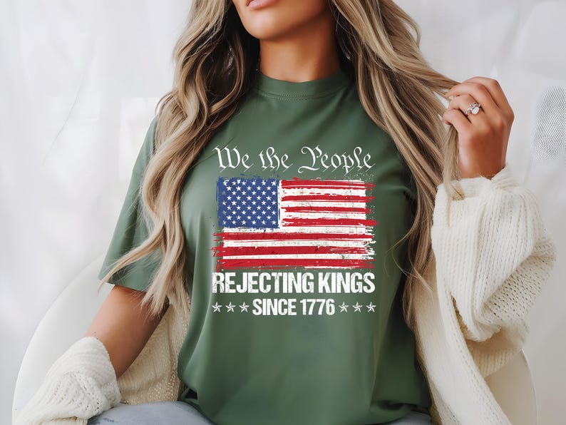 May include: Olive green t-shirt featuring a distressed American flag graphic with the text "We the People REJECTING KINGS SINCE 1776". The design includes red and white stripes, blue stars, and white text.