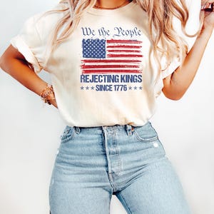 May include: Cream-colored t-shirt featuring a distressed American flag design in red, white, and blue. The text above the flag reads "We the People," and below it, "Rejecting Kings Since 1776." The shirt is paired with light blue jeans.