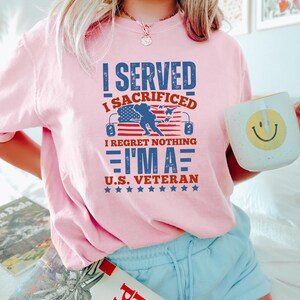 May include: A light pink t-shirt with the text "I SERVED I SACRIFICED I REGRET NOTHING I'M A U.S. VETERAN" in blue and red. The design includes an American flag and stars. The person is holding a white mug with a yellow smiley face.