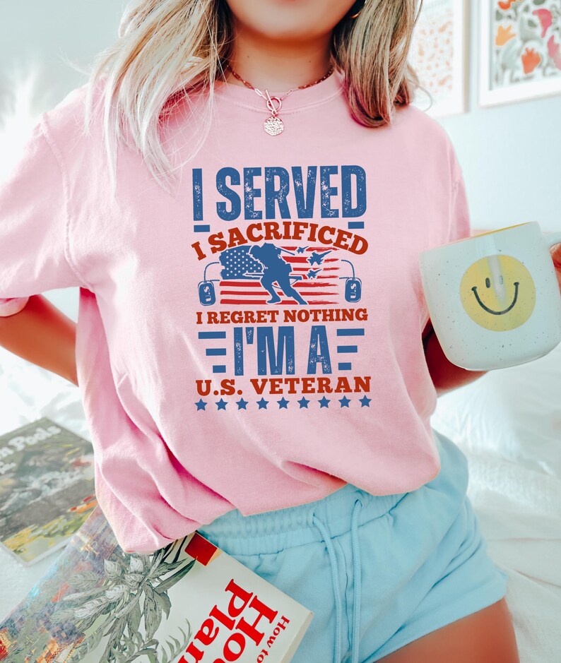 May include: A light pink t-shirt with the text "I SERVED I SACRIFICED I REGRET NOTHING I'M A U.S. VETERAN" in blue and red. The design includes an American flag and stars. The person is holding a white mug with a yellow smiley face.