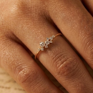 May include: A delicate gold ring featuring a cluster of small, sparkling diamonds. The ring is set on a slender band and is shown on a person's finger. The diamonds are clustered together, creating a floral or starburst effect.