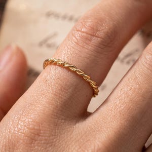 Op de afbeelding: Een dunne, goudkleurige ring met een delicate, gedraaide touwstructuur. De band heeft een gepolijste, metalen afwerking en een doorlopend, uniform ontwerp dat om de vinger zit, wat zorgt voor een eenvoudige en verfijnde uitstraling.