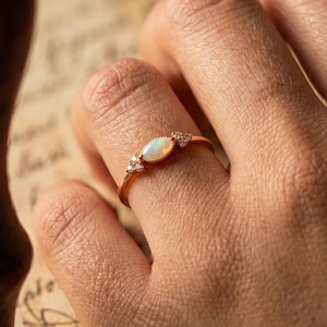 Könnte beinhalten: Ein roségoldener Ring mit einem ovalen Opal-Mittelstein und kleinen Akzentsteinen. Der Ring ist auf einem zarten Band gefasst. Der Opal hat ein milchig-weißes Aussehen. Der Ring wird am Finger gezeigt.