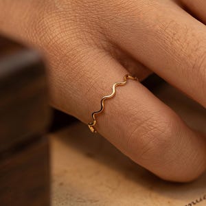 Può includere: Un anello color oro con un design a onda è indossato su un dito. L'anello è il punto focale, sullo sfondo di una mano, una scatola di legno e un pezzo di carta con testo manoscritto.