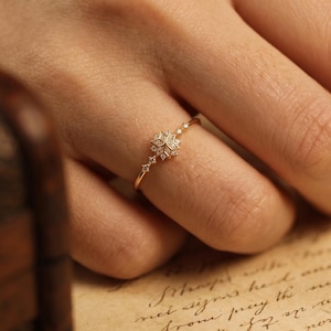 Può includere: Un delicato anello d'oro con un design a fiocco di neve e piccole gemme scintillanti. L'anello è indossato su un dito, mostrando i suoi dettagli intricati e il suo stile elegante. La fascia dell'anello è sottile.