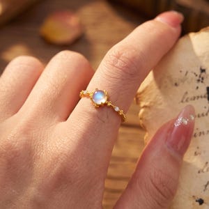 May include: A gold-toned ring featuring a central round iridescent moonstone cabochon, flanked by small clear stones on a delicate band with a textured, star-like metal setting that holds the stones in place on the finger.