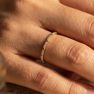 Könnte beinhalten: Ein goldener Ring mit kleinen, klaren Steinen, die um das Band herum gesetzt sind. Der Ring befindet sich an einem Finger, wobei die Hand auf einem Papier mit verblasstem Text ruht. Ein filigranes Schmuckstück.