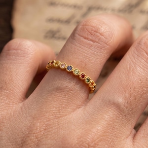 May include: A gold band ring featuring a series of small, round, multi-coloured gemstones set in a row. The metal band has a textured, beaded edge surrounding each individual stone, creating a continuous line of vibrant colours across the finger.