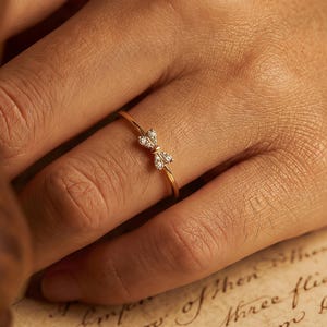 Könnte beinhalten: Ein goldener Ring mit einem schleifenförmigen Design, besetzt mit kleinen, klaren Edelsteinen. Der Ring befindet sich an einem Finger, mit einem unscharfen Hintergrund aus handschriftlichem Text auf gealtertem Papier. Das zarte Design des Rings ist der Fokus.