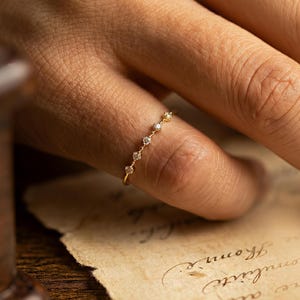 Könnte beinhalten: Ein zarter Goldring mit kleinen, funkelnden Edelsteinen entlang des Bandes. Der Ring wird an einem Finger präsentiert, der sein elegantes Design zeigt. Der schlichte, aber raffinierte Stil des Rings macht ihn zu einem vielseitigen Schmuckstück.