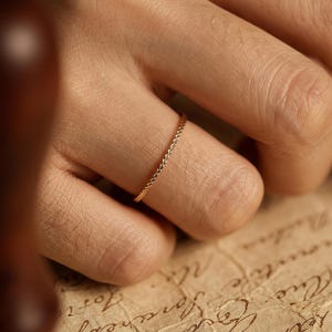 Könnte beinhalten: Ein zarter goldener Ring mit strukturiertem Design wird an einem Finger getragen. Der Ring ist dünn und scheint aus Gold zu sein. Der Hintergrund umfasst einen Holzstempel und ein Stück gealtertes Papier mit handschriftlichem Text.