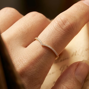 May include: A delicate, gold-toned ring with a curved design, adorned with small, sparkling gemstones. The ring is shown on a finger, highlighting its elegant and minimalist style. The ring's design is a subtle V-shape.