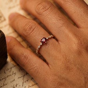 Puede incluir: Un anillo de oro con una piedra preciosa roja oscura en forma de corazón. El anillo tiene un diseño delicado con pequeñas piedras de acento claras y una banda detallada. El anillo está en el dedo de una persona.