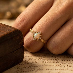 Könnte beinhalten: Ein goldener Ring mit einem ovalen Opal-Edelstein und kleinen, klaren Akzentsteinen. Der Ring hat ein zartes, verziertes Design mit einem filigranen Band. Der Ring wird an einem Finger getragen.