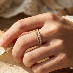 Puede incluir: Dos anillos de oro con piedras preciosas transparentes se muestran en un dedo. El anillo superior tiene una fila de piedras pequeñas, mientras que el anillo inferior presenta piedras más grandes y espaciadas. El fondo es suave y borroso.