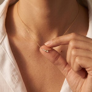 Può includere: Una delicata collana a catena dorata con un ciondolo a barra orizzontale incastonato con piccole pietre chiare e scintillanti. Il ciondolo è fissato a una sottile catena dorata lucida che poggia sul collo, mostrando un design raffinato e semplice.