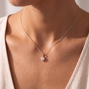 May include: A gold chain necklace featuring a single round, clear, faceted gemstone pendant held in a four-prong setting. The delicate metal chain rests against the neck, showcasing the brilliant cut of the stone which reflects light.