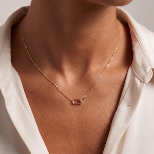 May include: A gold-toned necklace featuring a delicate chain and a small, rectangular link pendant. The pendant consists of two interlocking rectangular loops, with one loop appearing slightly thicker than the other, resting centrally on the chain.