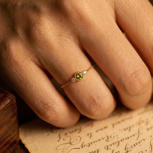 Op de afbeelding: Een delicate gouden ring met een centrale, ronde, lichtgroene edelsteen, geflankeerd door twee kleinere witte edelstenen. De ring is gezet op een dunne gouden kettingband, wat een minimalistisch en elegant ontwerp creëert. De ring wordt op een vinger getoond.
