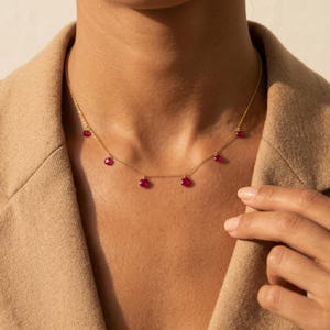 Op de afbeelding: Een delicate gouden ketting met vijf gelijkmatig verdeelde, kleine, ronde, gefacetteerde dieproze edelstenen die als individuele bedels langs de ketting hangen en rusten tegen de basis van de nek.
