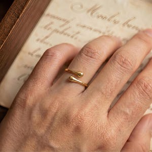 May include: A gold-toned open ring featuring two smooth, rounded teardrop-shaped ends that face each other. The band is polished and reflective, creating a continuous loop design that remains open at the center of the finger.