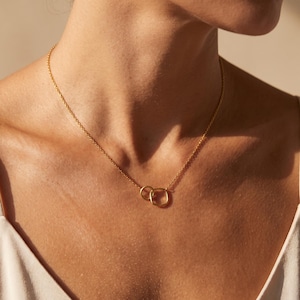 May include: A gold-toned necklace featuring a delicate chain and a pendant composed of two interlocking geometric shapes, one circular and one square, resting against the collarbone with a polished metallic finish and a simple clasp closure.