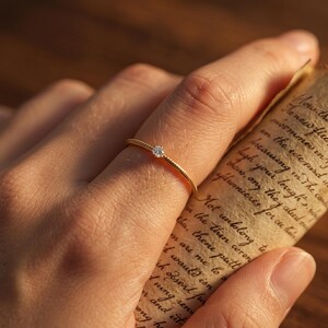 May include: A thin gold ring featuring a single small, clear, round-cut gemstone set in the centre. The band of the ring has a delicate, textured, beaded surface, providing a subtle detail to the minimalist design of this piece of jewellery.