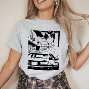 May include: A light gray t-shirt featuring a black and white anime-style graphic. The graphic depicts a car, a person, and Japanese text. The car has the number "13-600" on its license plate.