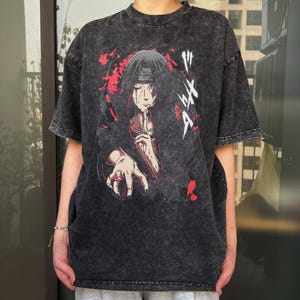 May include: Dark gray, oversized t-shirt featuring a graphic of a character with red and black accents. The character is depicted with a hand raised to the mouth and a ring on the finger. Japanese text is visible on the right side of the graphic.