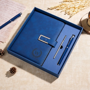 Könnte beinhalten: Ein blaues Notizbuch mit einem goldfarbenen rechteckigen Verschluss und einem passenden blauen Stift in einer blauen Geschenkbox. Der Einband des Notizbuchs zeigt ein Lorbeerkranz-Emblem um den Buchstaben W und den Namen William Brown.