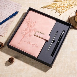 May include: A pink notebook with a floral line art design and the name Amelia engraved on the cover, featuring a gold-toned metal clasp. The notebook is presented in a black gift box alongside a black ballpoint pen and a silver stylus.