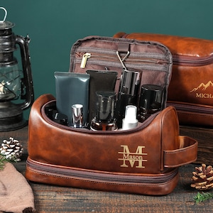 May include: A brown leather toiletry bag with a zippered main compartment and a secondary bottom compartment. The front features the name Mason in a stylized monogram. The bag is open, revealing various dark-colored cosmetic bottles inside.