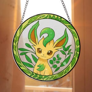 以下が含まれることがあります： 黄色と緑色の漫画のキャラクターが描かれた丸いステンドグラスパネル。緑の葉に囲まれています。 パネルは緑色の縁取りがあり、チェーンから吊り下げられています。