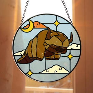 Puede incluir: Vidriera redonda que representa una criatura marrón con forma de bisonte con cuernos, sobre un cielo azul con nubes, una luna creciente y estrellas. La obra está enmarcada con un borde de metal plateado y colgada de una cadena.