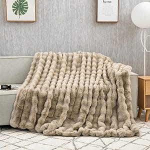 May include: A beige faux fur throw blanket with a textured, vertical ribbed pattern. The blanket is draped over a light gray sofa, featuring a soft, plush appearance with distinct raised ridges running across the entire surface of the fabric.