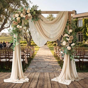May include: A wooden wedding arch draped with sheer beige fabric and decorated with arrangements of cream, tan, and brown artificial flowers mixed with green eucalyptus leaves, positioned on a wooden platform outdoors.