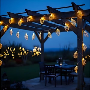 May include: Outdoor scene at dusk featuring a wooden pergola adorned with string lights. The lights are shaped like golden leaves, casting a warm glow. A table and chairs are visible, suggesting a patio setting, perfect for evening gatherings.