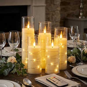 May include: Five cylindrical glass vases containing cream-colored LED pillar candles wrapped in thin, glowing fairy light strings. The candles are arranged on a table with wine glasses, white plates, greenery, and a small remote control.